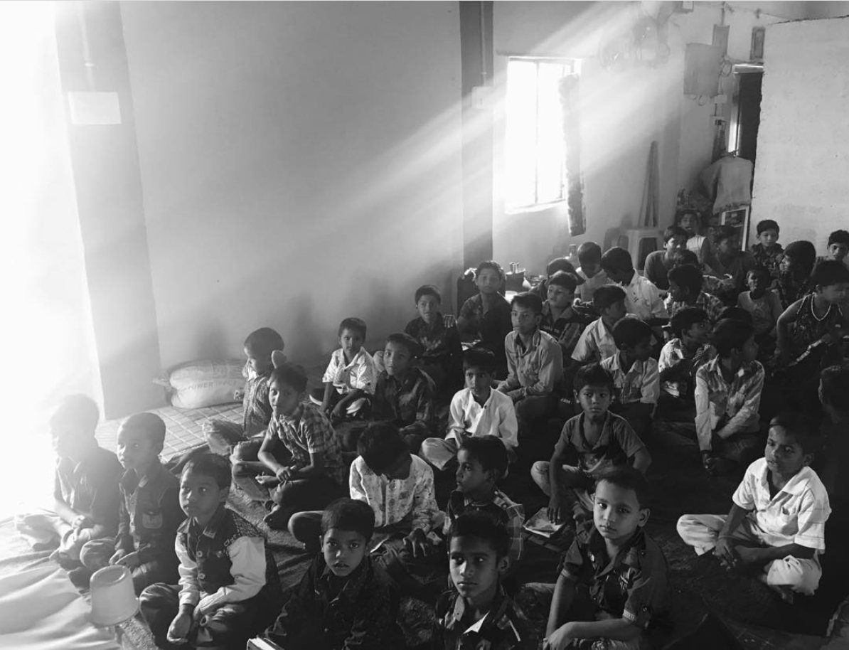 Children sitting on the floor of a mission classroom, lit by a single window
