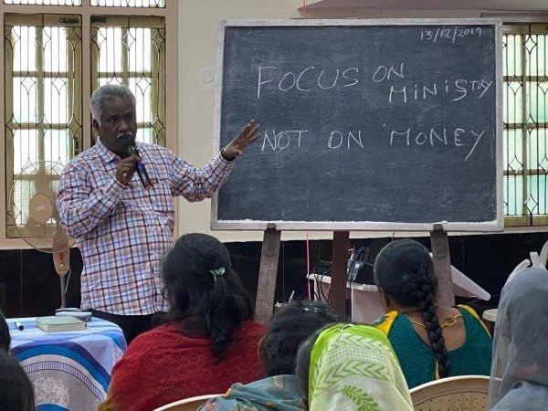 A pastor teaching a group, with “focus on ministry, not on money” written on the board behind him